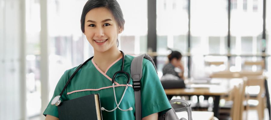 Eine junge asiatische Pflegekraft steht in einem Lehrraum, hält ein Buch in der Hand und schaut lächelnd in die Kamera