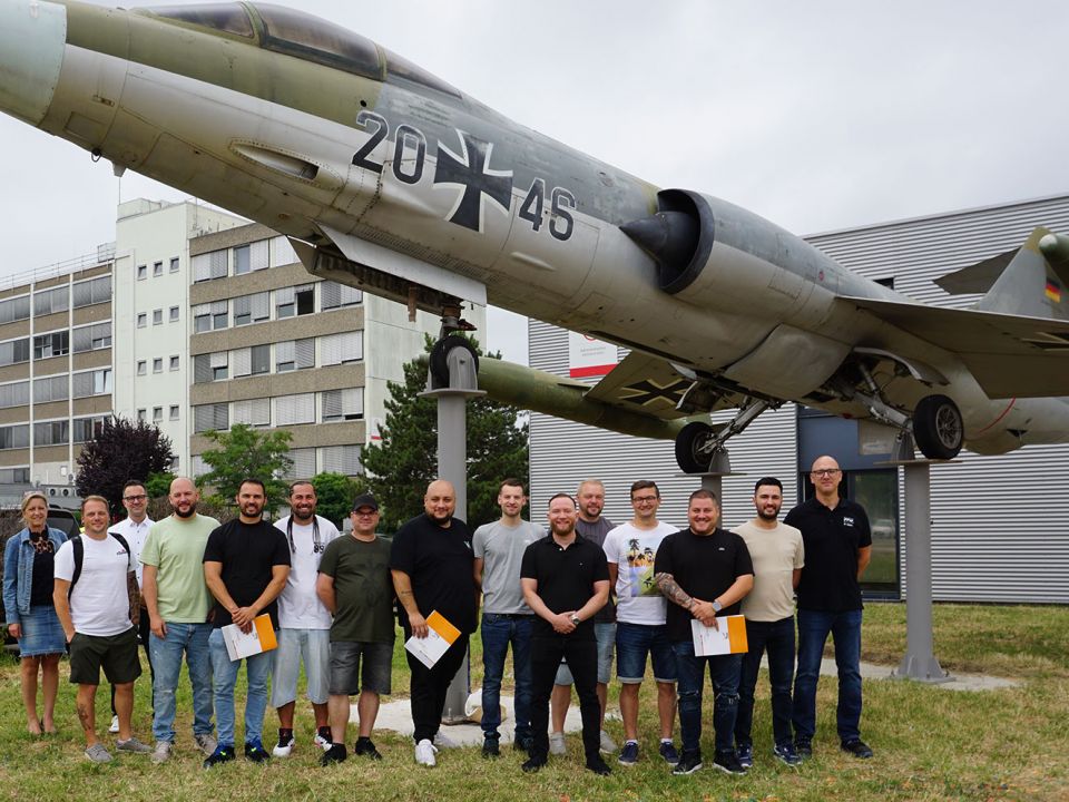Die Absolventen der Umschulung Industriemechaniker stehen unter einem Militärflugzeug auf dem Gelände der PFW Aerospace GmbH für ein Gruppenfoto zusammen.
