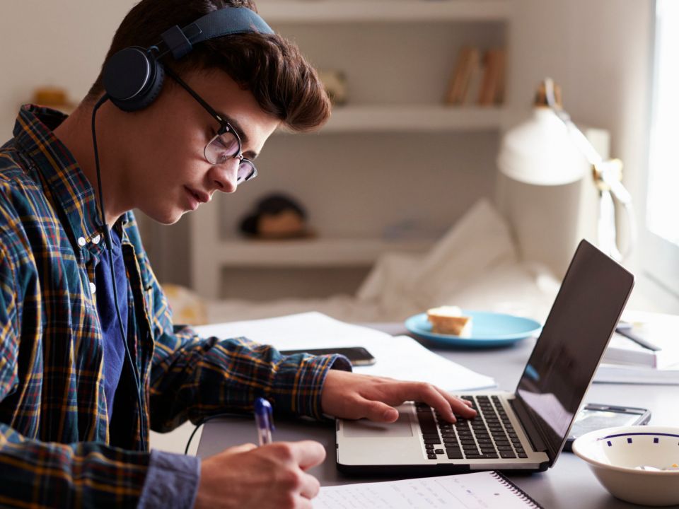 Ein Schüler sitzt an seinem Schreibtisch, arbeitet mit Kopfhörern an seinem Laptop und macht sich Notizen
